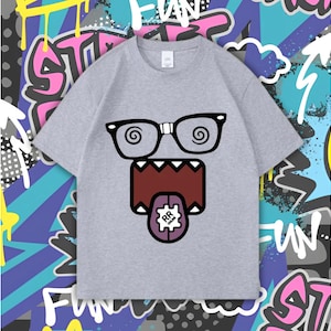 Op de afbeelding: Lichtgrijs t-shirt met een cartoongezicht met een bril, een wijd open mond en een tong met een tandradpatroon. De achtergrond is een kleurrijk graffiti-ontwerp.