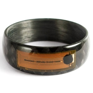 Puede incluir: Un anillo negro de fibra de carbono con un patrón geométrico. El anillo presenta una incrustación rectangular de color cobre con un punto negro. Es un accesorio tecnológico portátil.