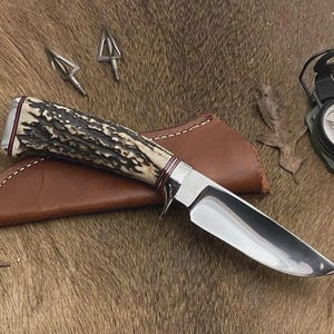 May include: A hunting knife with a polished steel blade and a handle made of antler material. The knife rests on a brown leather sheath, with two arrowheads and a compass nearby. The knife is a hunting tool.