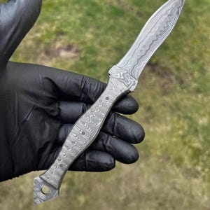May include: A Damascus steel knife with a patterned blade and handle. The knife has a pointed tip and a handle with a series of small, round indentations. The blade and handle have a swirling, textured appearance.