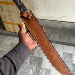 May include: A long, sharp knife with a dark wooden handle and a brown leather sheath. The sheath has a decorative pattern and a snap closure. The knife is held in a person's hand.