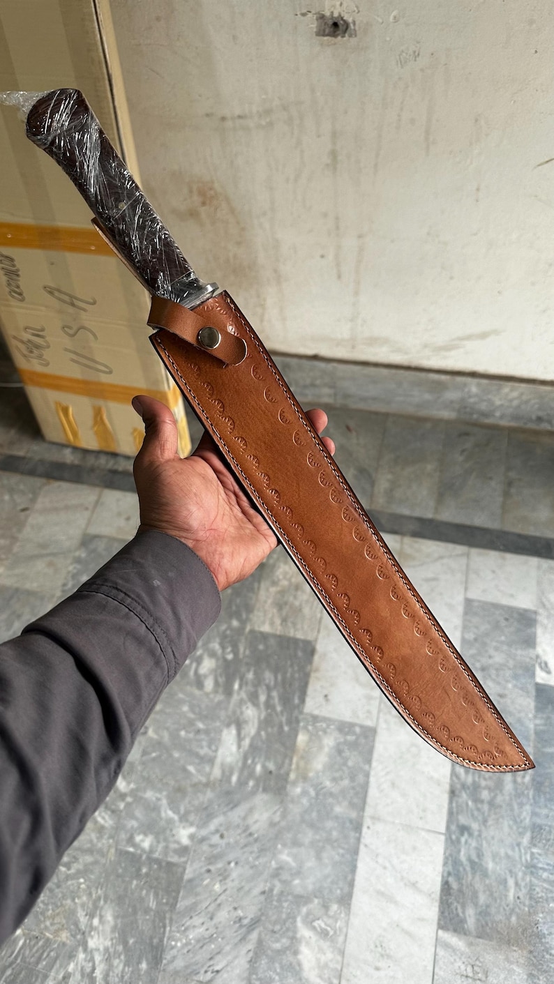 May include: A long, sharp knife with a dark wooden handle and a brown leather sheath. The sheath has a decorative pattern and a snap closure. The knife is held in a person's hand.