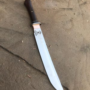 May include: A handmade knife with a polished steel blade and a dark wood handle. The handle is attached to the blade with a copper bolster. The blade has etched text and a symbol. The knife is displayed on a brown surface.