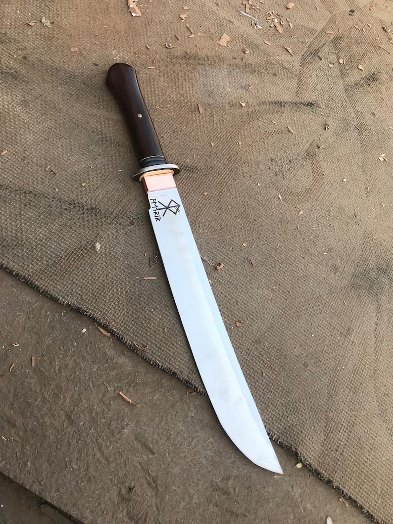 May include: A handmade knife with a polished steel blade and a dark wood handle. The handle is attached to the blade with a copper bolster. The blade has etched text and a symbol. The knife is displayed on a brown surface.