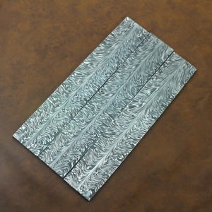 Puede incluir: Cinco piezas rectangulares de acero con un patrón arremolinado único en tonos grises y blancos. El patrón se asemeja al agua que fluye o a un efecto veteado. Las piezas de acero están dispuestas diagonalmente sobre una superficie marrón.