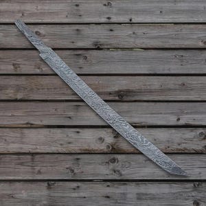 Puede incluir: Una larga hoja de espada plateada con un diseño estampado. La hoja es de metal y tiene un filo afilado. La espada está sobre una superficie de madera.