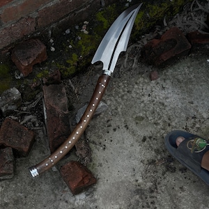 May include: A curved spear with a polished silver-toned metal blade featuring a sharp point and a notched base. The handle is made of dark brown wood, accented with a single row of small, circular silver-toned metal inlays along its length.