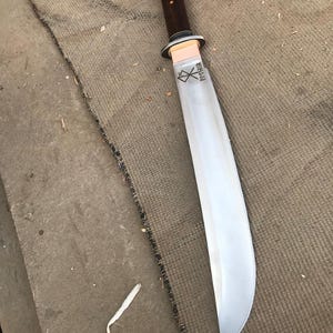 May include: A large, curved blade knife with a dark wooden handle and a polished steel blade. The handle is attached to the blade with a copper-colored guard. The blade has etched text near the handle.