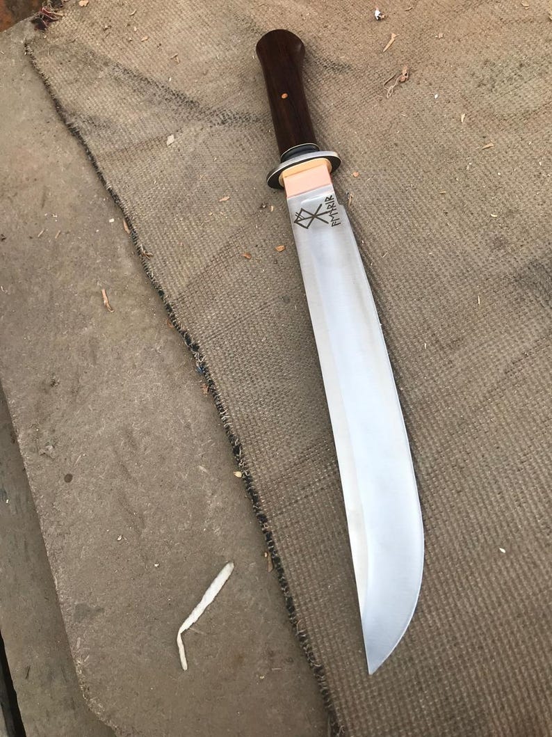 May include: A large, curved blade knife with a dark wooden handle and a polished steel blade. The handle is attached to the blade with a copper-colored guard. The blade has etched text near the handle.