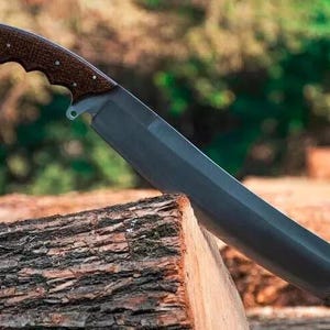 May include: A large, dark-bladed knife with a brown, textured handle. The knife is angled, resting on a piece of wood, with a blurred natural background. The handle has a curved design for grip.