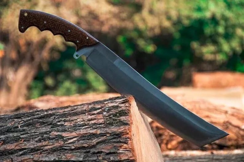 May include: A large, dark-bladed knife with a brown, textured handle. The knife is angled, resting on a piece of wood, with a blurred natural background. The handle has a curved design for grip.