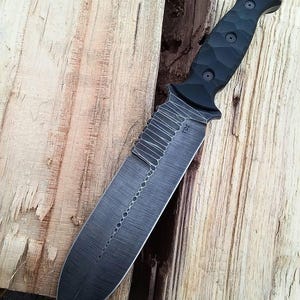 May include: A black tactical knife with a textured handle and a partially serrated blade. The blade has a series of small, circular cutouts along its length. The knife rests on a wooden surface.