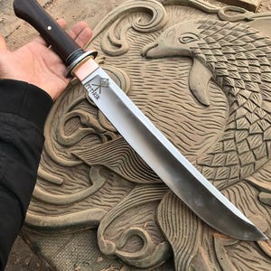 May include: A handcrafted knife with a dark wooden handle and a silver blade. The blade has the word "FMRIR" etched on it, along with a symbol. The knife is held over a carved wooden surface.