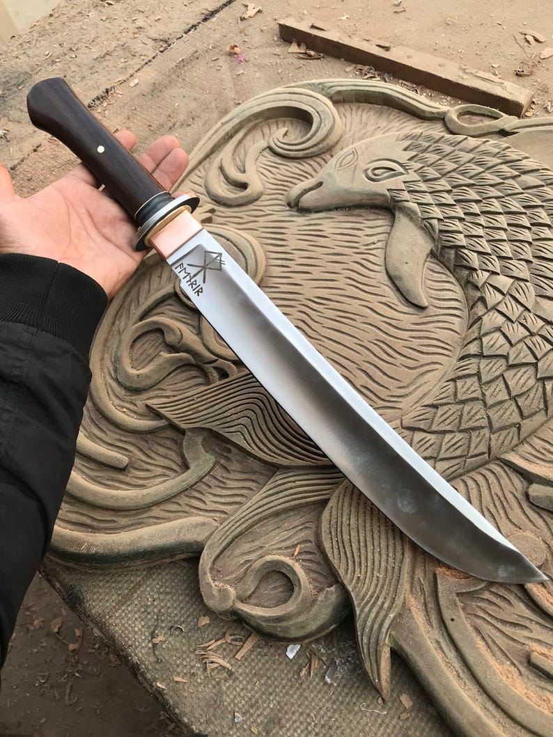 May include: A handcrafted knife with a dark wooden handle and a silver blade. The blade has the word "FMRIR" etched on it, along with a symbol. The knife is held over a carved wooden surface.