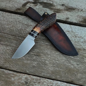 Puede incluir: Un cuchillo de caza artesanal con una hoja de acero pulido y un mango de material marrón oscuro texturizado. El cuchillo descansa sobre una superficie de madera junto a una funda de cuero marrón.