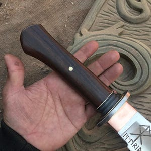 May include: A handcrafted knife with a dark wood handle and a silver blade. The blade has etched designs and the word "Fyrir" is visible. The handle is dark brown with a small white dot. The knife is held in a hand.