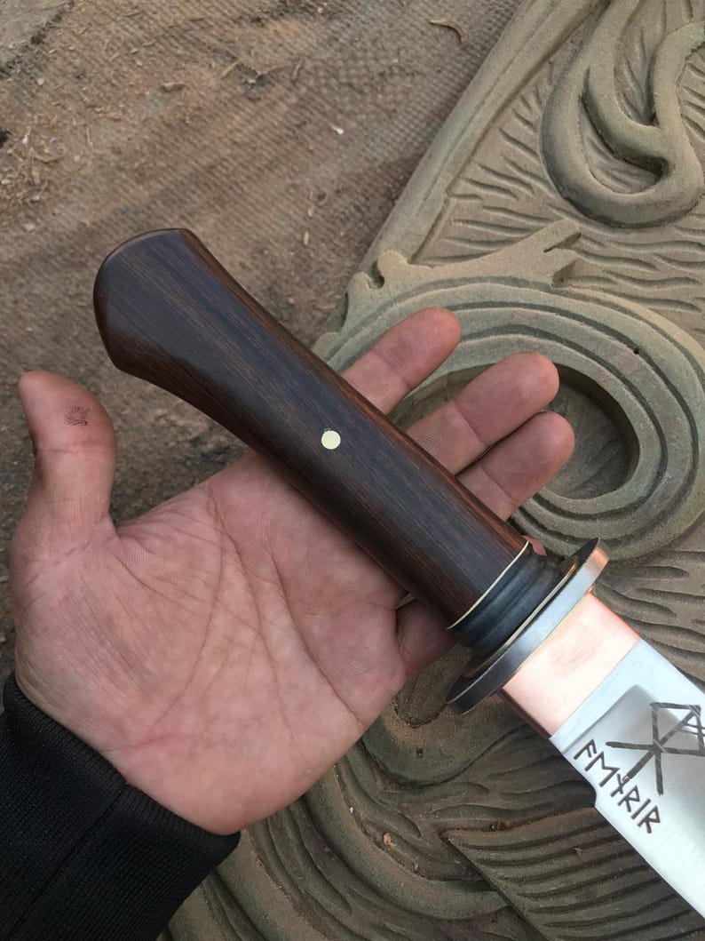 May include: A handcrafted knife with a dark wood handle and a silver blade. The blade has etched designs and the word "Fyrir" is visible. The handle is dark brown with a small white dot. The knife is held in a hand.