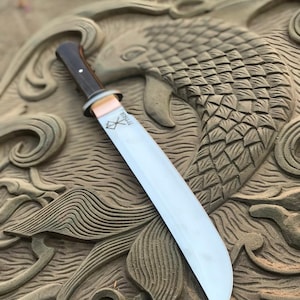 May include: A large, polished steel blade knife with a dark wooden handle and a copper-colored bolster. The blade has a slight curve and a sharp edge. The knife rests on a carved stone surface with a fish design.