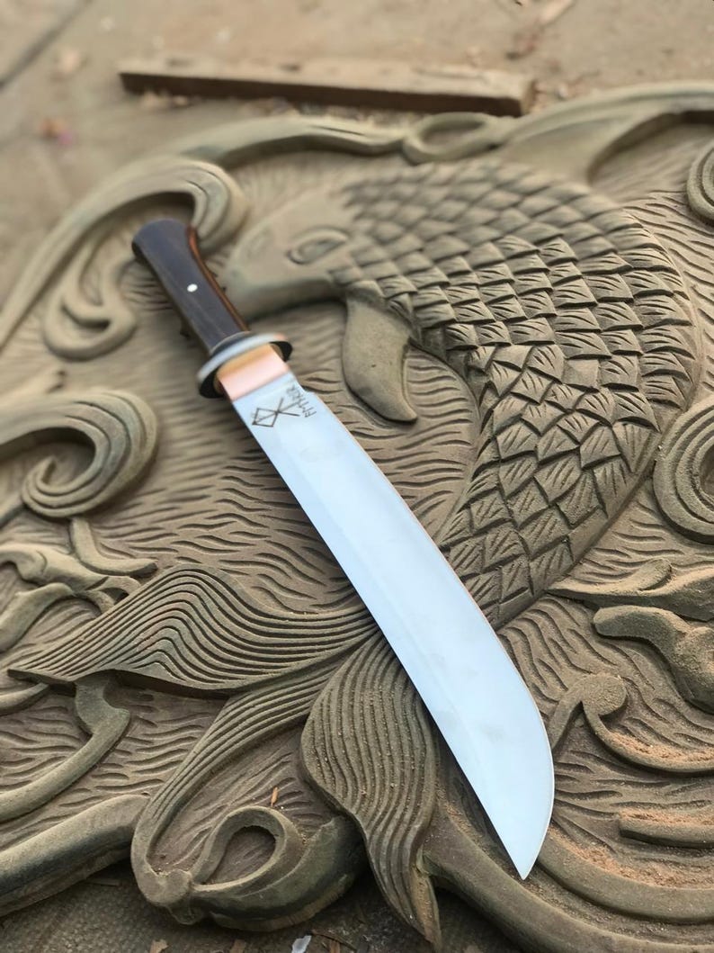 May include: A large, polished steel blade knife with a dark wooden handle and a copper-colored bolster. The blade has a slight curve and a sharp edge. The knife rests on a carved stone surface with a fish design.