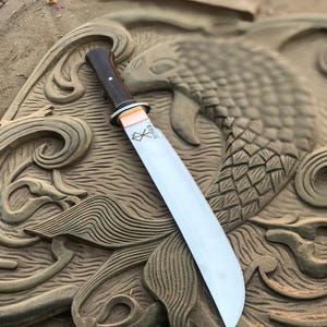 May include: A large, polished steel knife with a dark wooden handle and a copper bolster. The blade is engraved with the word "THOR" and a symbol. The knife is set against a carved wooden background.