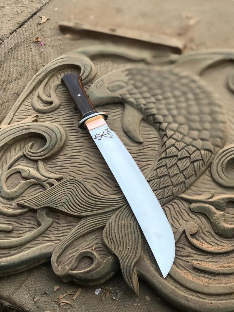 May include: A large, polished steel knife with a dark wooden handle and a copper bolster. The blade is engraved with the word "THOR" and a symbol. The knife is set against a carved wooden background.