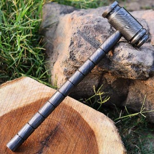 May include: A detailed, dark gray hammer with a patterned head and handle. The hammer is resting on a wooden surface and a large rock. The hammer's handle is approximately 12 inches long.