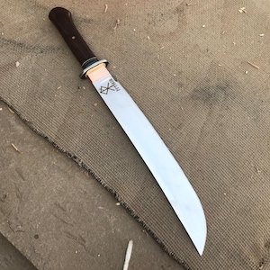 May include: A long, silver blade knife with a dark brown wooden handle. The blade has the word "Mimir" etched on it. The knife is lying on a brown, textured surface.