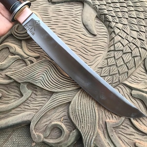 May include: A handcrafted knife with a dark wooden handle, a silver blade, and a copper bolster. The blade has etched text. The knife is displayed on a carved wooden surface with a fish design.