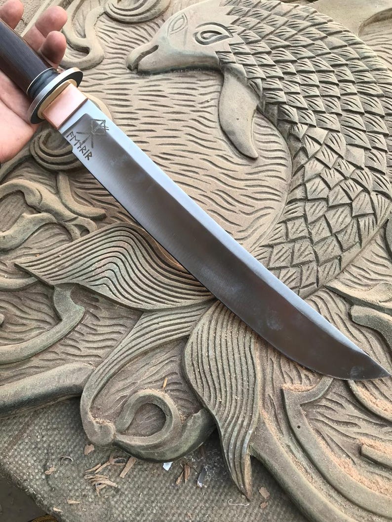 May include: A handcrafted knife with a dark wooden handle, a silver blade, and a copper bolster. The blade has etched text. The knife is displayed on a carved wooden surface with a fish design.