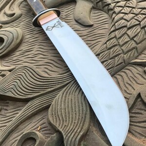 May include: A large, curved blade knife with a wooden handle and a copper bolster. The blade is silver and has etched markings. The knife rests on a carved wooden surface with a fish or dragon design.