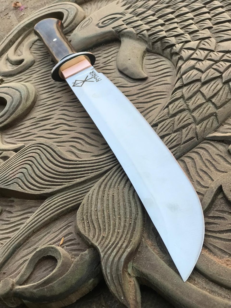 May include: A large, curved blade knife with a wooden handle and a copper bolster. The blade is silver and has etched markings. The knife rests on a carved wooden surface with a fish or dragon design.