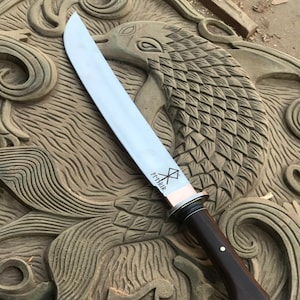 May include: A large, polished steel knife with a dark wooden handle and a copper bolster. The blade has etched markings. The knife rests on a carved wooden surface with a bird design.