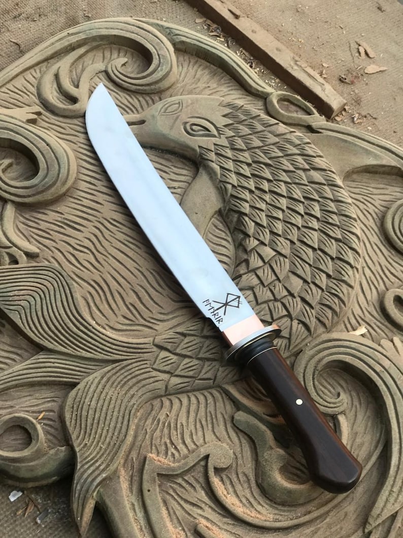May include: A large, polished steel knife with a dark wooden handle and a copper bolster. The blade has etched markings. The knife rests on a carved wooden surface with a bird design.