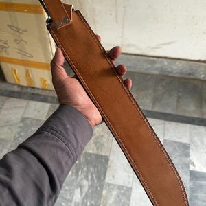 May include: A large, brown leather sheath with a knife inside. The sheath is stitched with white thread and has a strap with two silver rivets. The knife handle is visible, and the blade is partially concealed. The knife is held by a person.