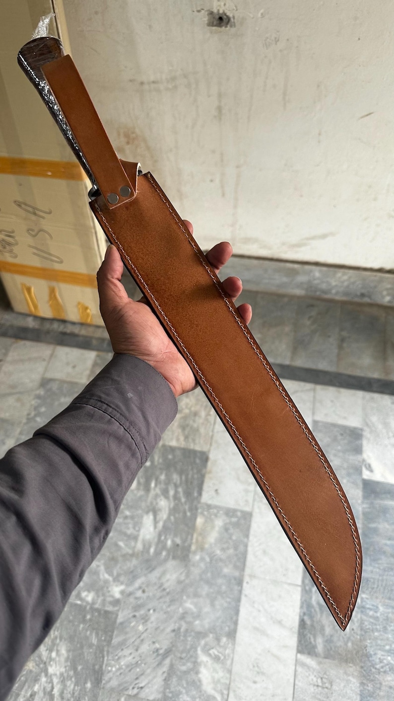 May include: A large, brown leather sheath with a knife inside. The sheath is stitched with white thread and has a strap with two silver rivets. The knife handle is visible, and the blade is partially concealed. The knife is held by a person.