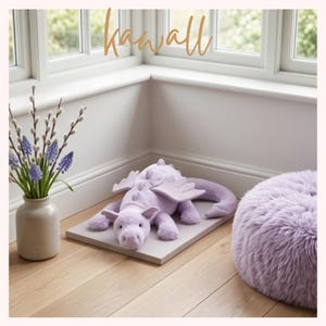 Puede incluir: Un peluche de dragón lavanda con alas y una larga cola descansa sobre una plataforma gris. Un puf difuso a juego y un jarrón con flores moradas están cerca. La palabra "kawaii" está escrita arriba.