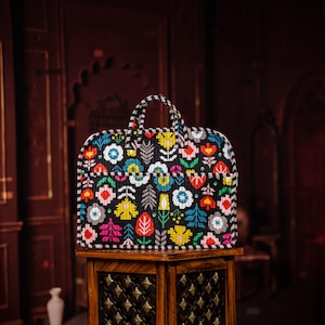 Puede incluir: Un bolso tote negro con un colorido estampado floral. El bolso presenta una variedad de flores en tonos rojos, rosas, amarillos y azules. Tiene un asa a rayas y está sobre una mesa de madera.