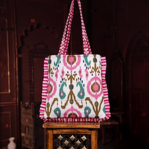 Puede incluir: Un bolso tote blanco con un colorido estampado ikat en rosa, turquesa y marrón. El bolso tiene asas a rayas rosas y blancas y un borde a rayas rosas. El bolso está sobre una pequeña mesa de madera.