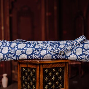 May include: A rolled-up yoga mat with a blue and white floral pattern. The mat is made of a quilted fabric and has a strap for carrying. The mat is resting on a wooden table with decorative metal accents. The background is a dark, blurred interior.