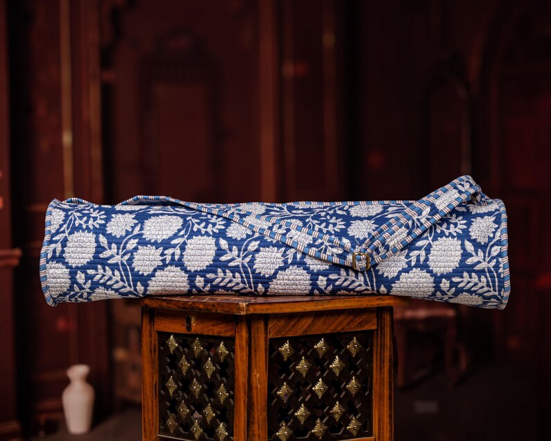 May include: A rolled-up yoga mat with a blue and white floral pattern. The mat is made of a quilted fabric and has a strap for carrying. The mat is resting on a wooden table with decorative metal accents. The background is a dark, blurred interior.