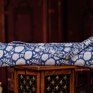 May include: Rolled-up blue and white floral patterned yoga mat. The mat is made of a quilted fabric and has a strap with a gold-colored buckle. The mat is resting on a small wooden table.