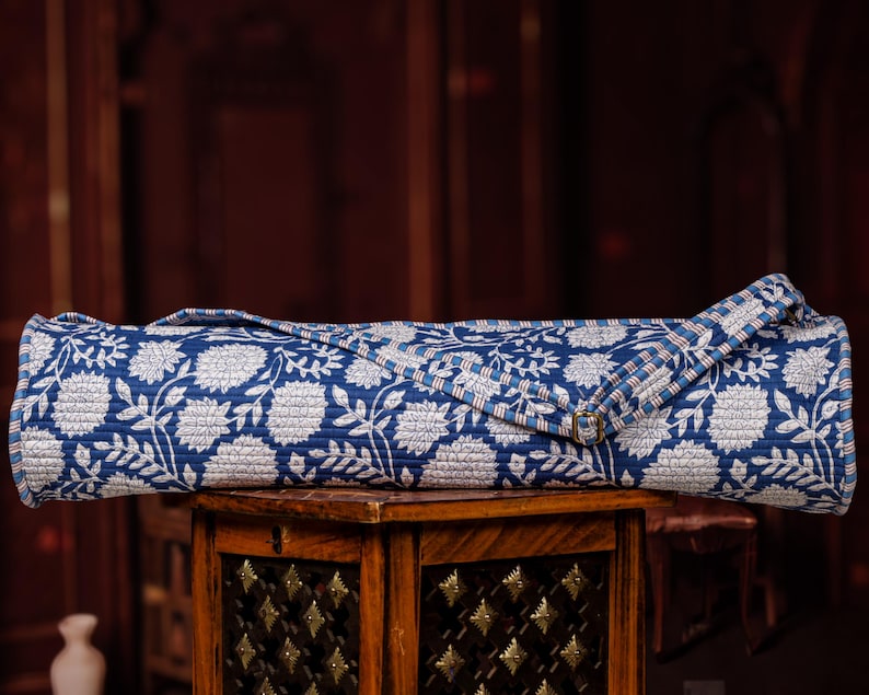 May include: Rolled-up blue and white floral patterned yoga mat. The mat is made of a quilted fabric and has a strap with a gold-colored buckle. The mat is resting on a small wooden table.