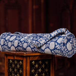 May include: A rolled-up, quilted yoga mat bag with a blue and white floral pattern. The bag has a handle and is resting on a wooden table. The bag's design features a repeating pattern of white flowers and leaves on a blue background.