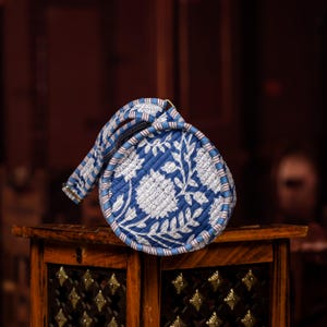May include: A round, blue and white floral patterned clutch with a matching handle. The clutch is made of quilted fabric and features a zipper closure. The design includes white floral motifs on a blue background.