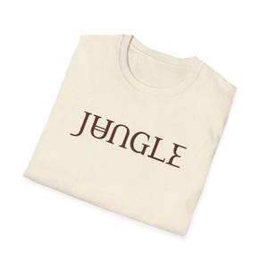 May include: Cream-colored t-shirt with the word "JUNGLE" printed in a dark brown, stylized font. The shirt is folded and appears to be made of a soft, comfortable material. The design is simple and eye-catching.