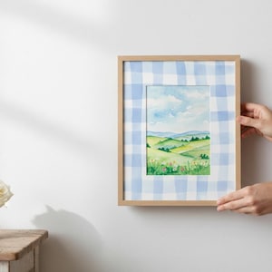 May include: A framed watercolor landscape painting featuring rolling green hills, a blue sky with clouds, and wildflowers. The frame is light wood, and the matting has a blue and white gingham pattern.