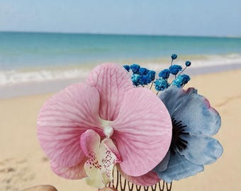 May include: A hair comb featuring a large pink orchid with a white and speckled center, a blue fabric flower with a dark center, and small clusters of bright blue dried flowers attached to a brown metal comb base.