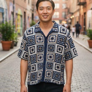 May include: A short-sleeved, button-down shirt with a crochet pattern in shades of blue, gray, and cream. The shirt has a collar and is worn with black pants. The shirt's design features square motifs.