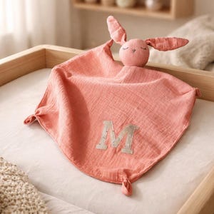 Könnte beinhalten: Eine korallenfarbene Baby-Schmusedecke mit einem Plüsch-Hasenkopf und Ohren. Die Decke hat den Buchstaben "M" in einer neutralen Farbe appliziert. Die Ecken sind geknotet. Ein weicher und tröstender Artikel für Babys.