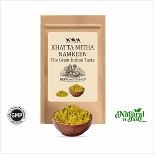 Pode incluir: Um saco de papel castanho com uma etiqueta que diz "KHATTA MITHA NAMKEEN The Great Indian Taste". Uma tigela de madeira é preenchida com uma mistura de lanches amarelo-verde. As palavras "Natural n Tasty" são verdes.
